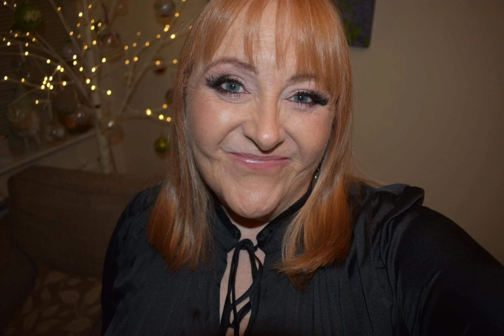 A woman with shoulder-length reddish hair taking a selfie indoors, with a decorated Christmas tree and warm lighting in the background.