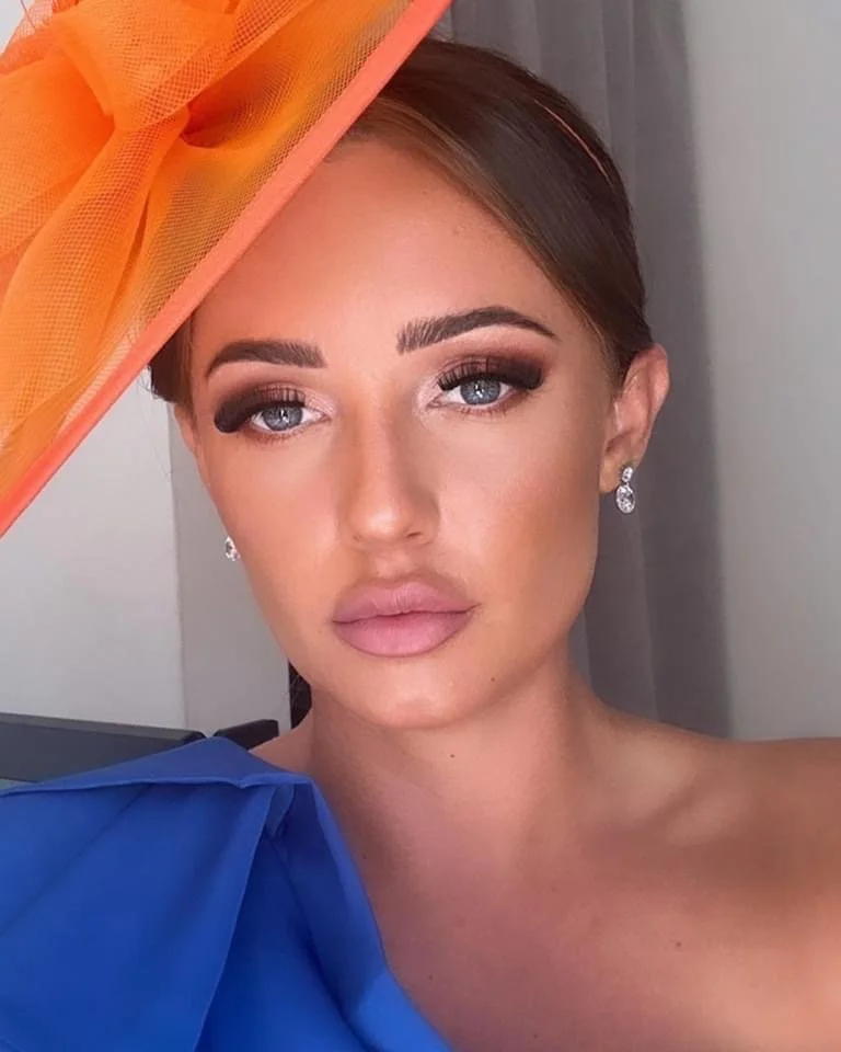 A woman with makeup and earrings wearing a large draped garment, standing indoors with a gray curtain background and some orange and blue fabric.