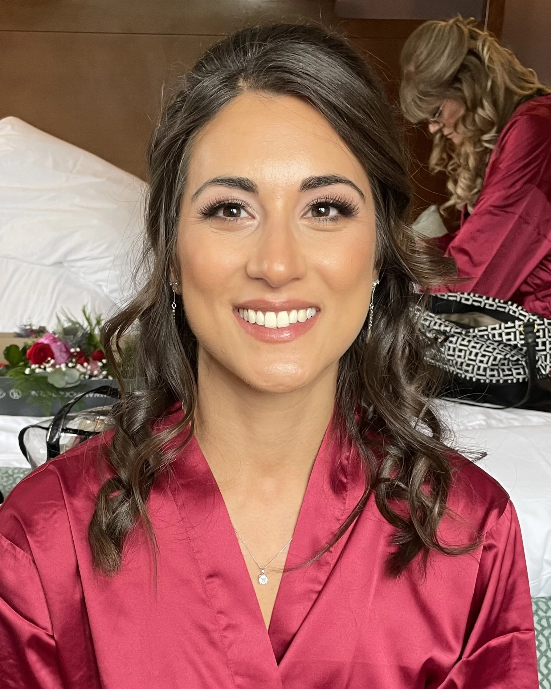 A woman with curly brown hair, wearing a red outfit and jewelry, is smiling at the camera. In the background, another woman with blonde hair, glasses, and a red jacket is working or preparing something. There is a bouquet of flowers on the table in f