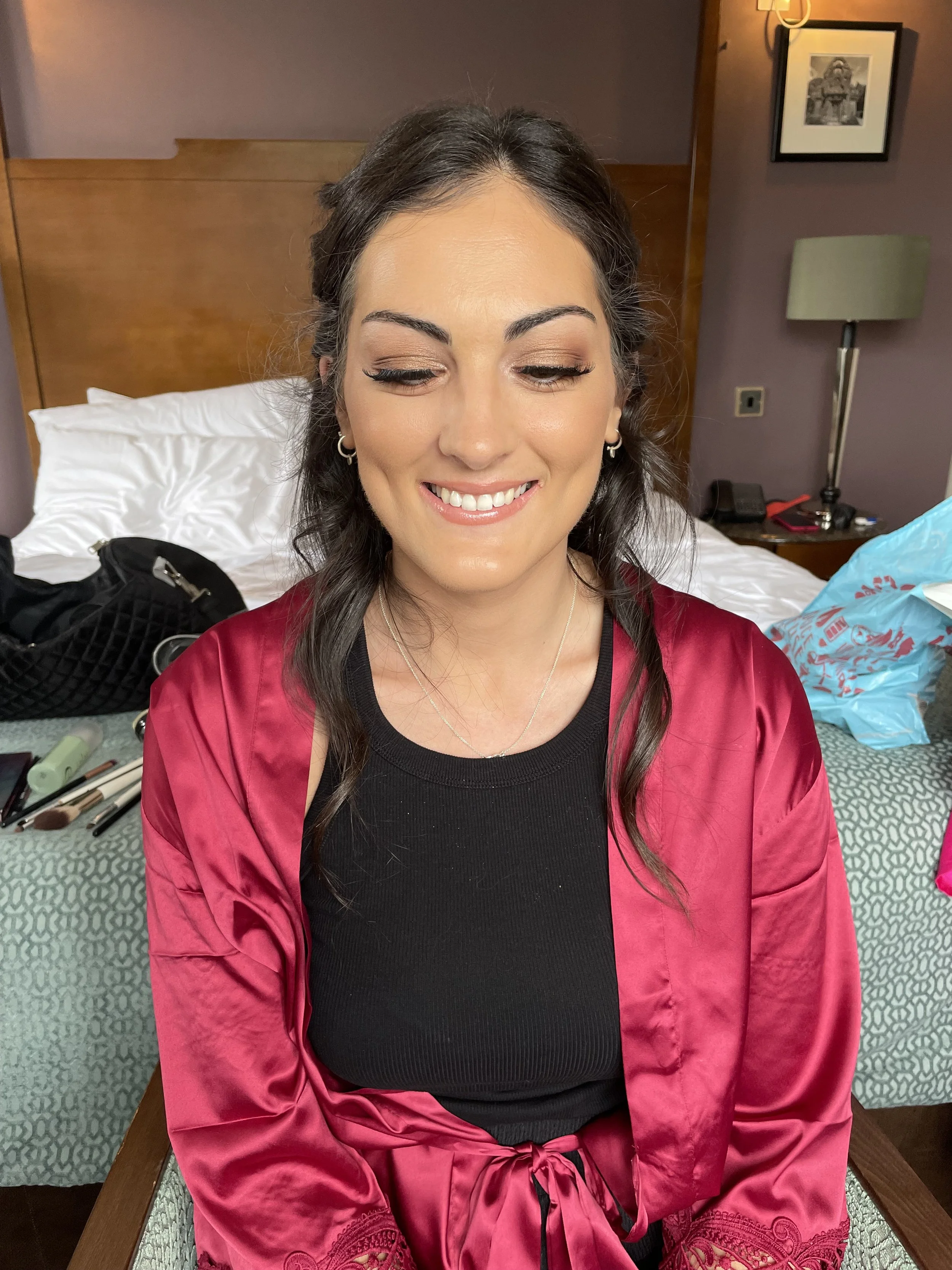 A woman with dark hair, styled in loose waves, smiling with her eyes closed, wearing makeup including eye shadow, eyeliner, and lipstick, dressed in a black top and a pink satin robe with embroidery, sitting in a hotel room with a bed, makeup tools, 