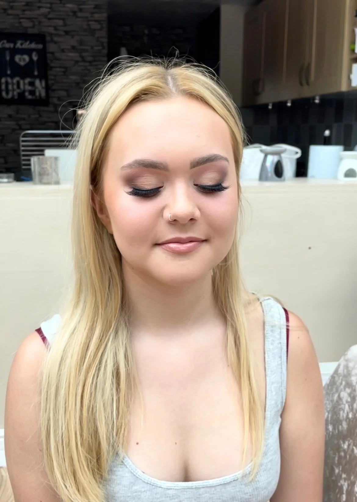 A woman with blonde hair and closed eyes, wearing makeup, sitting in a kitchen.