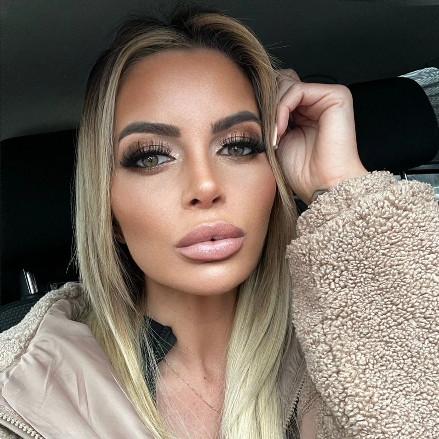 A woman with blonde hair and makeup, wearing a beige jacket with a textured, fleece-like sleeve, sitting in a car and resting her hand on her head.