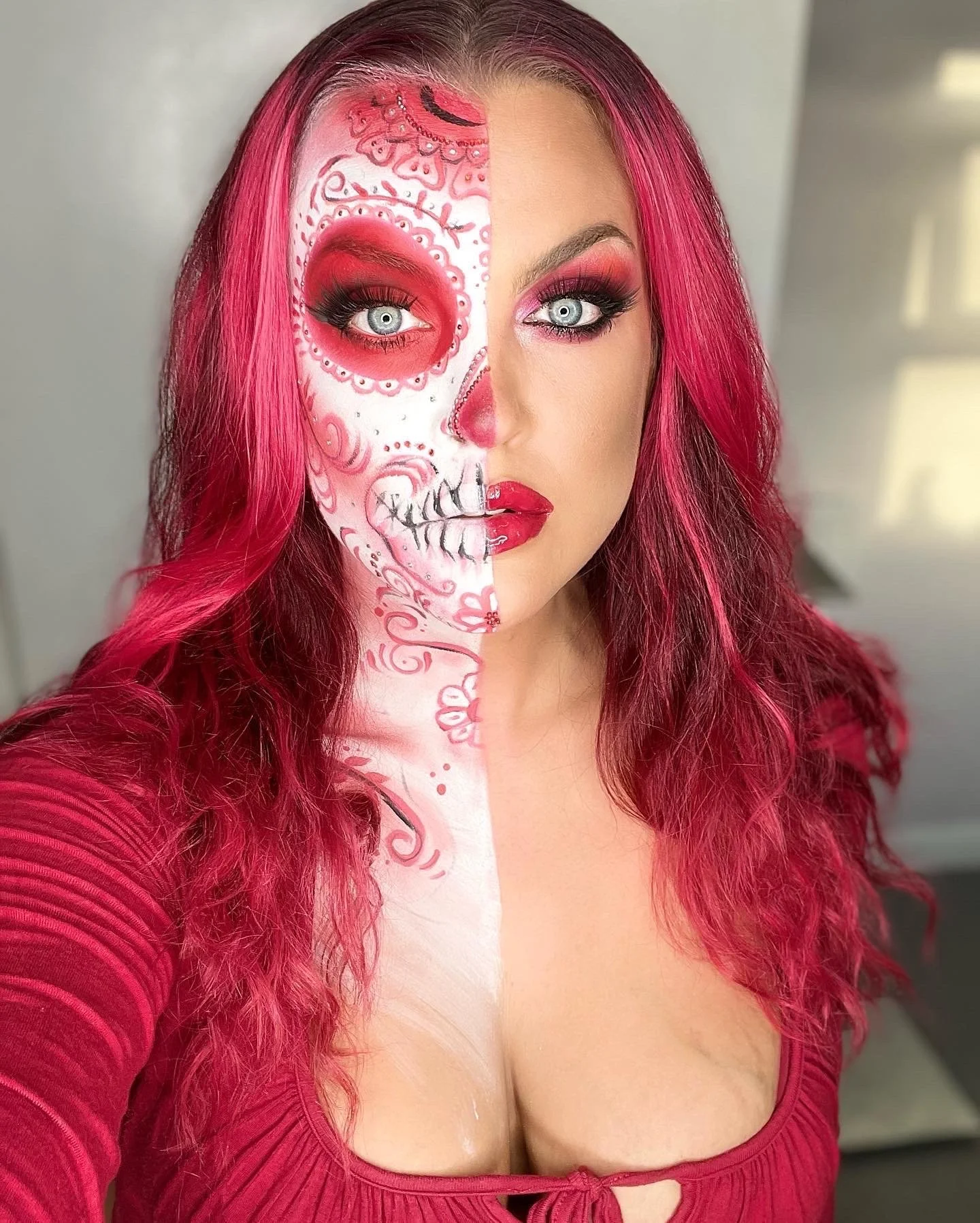 A woman with long, curly pink hair taking a selfie. Half of her face is painted like a decorative sugar skull with pink and white details, and the other half shows her makeup with pink eyeshadow, black eyeliner, and red lipstick. She is wearing a red