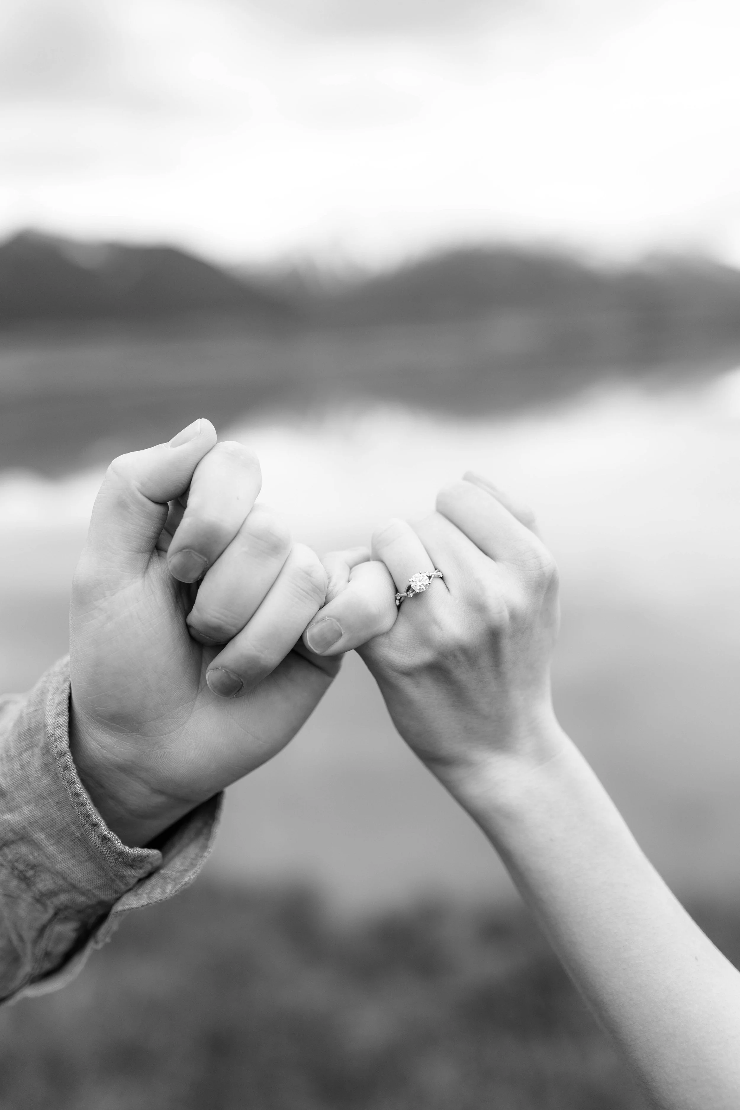 Two hands holding each other, one with an engagement ring on the ring finger, against a blurred outdoor background.