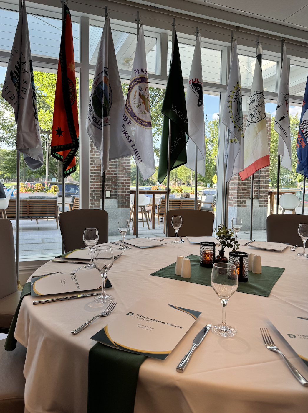 Round banquet table set for a meeting or event with documents, wine glasses, and candles, positioned near large windows with flags hanging inside the room, and outdoor seating with trees visible outside.