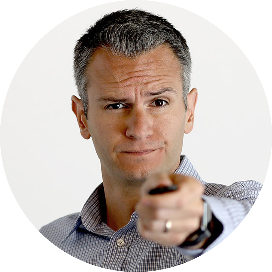 A man with short, graying hair is pointing and looking directly at the camera with a slightly raised eyebrow and smirk.