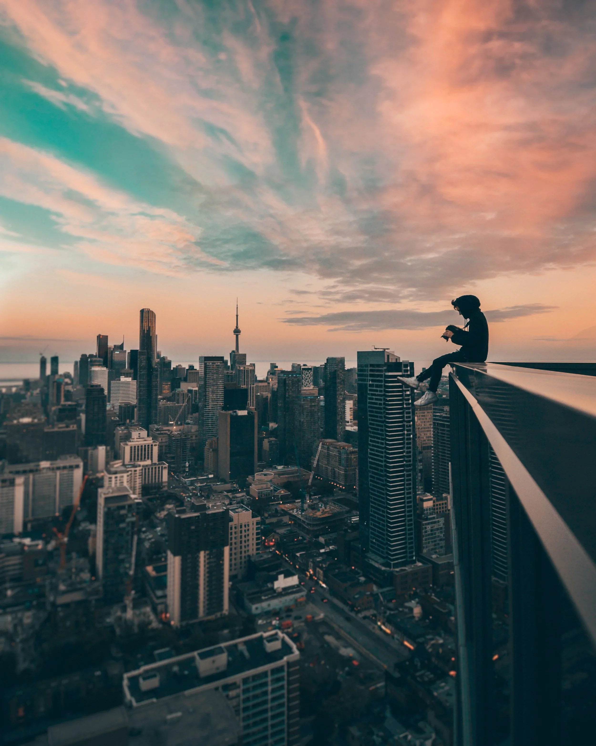 Silhouette of a person sitting on the edge of a rooftop with city skyline, including CN Tower, during sunset.