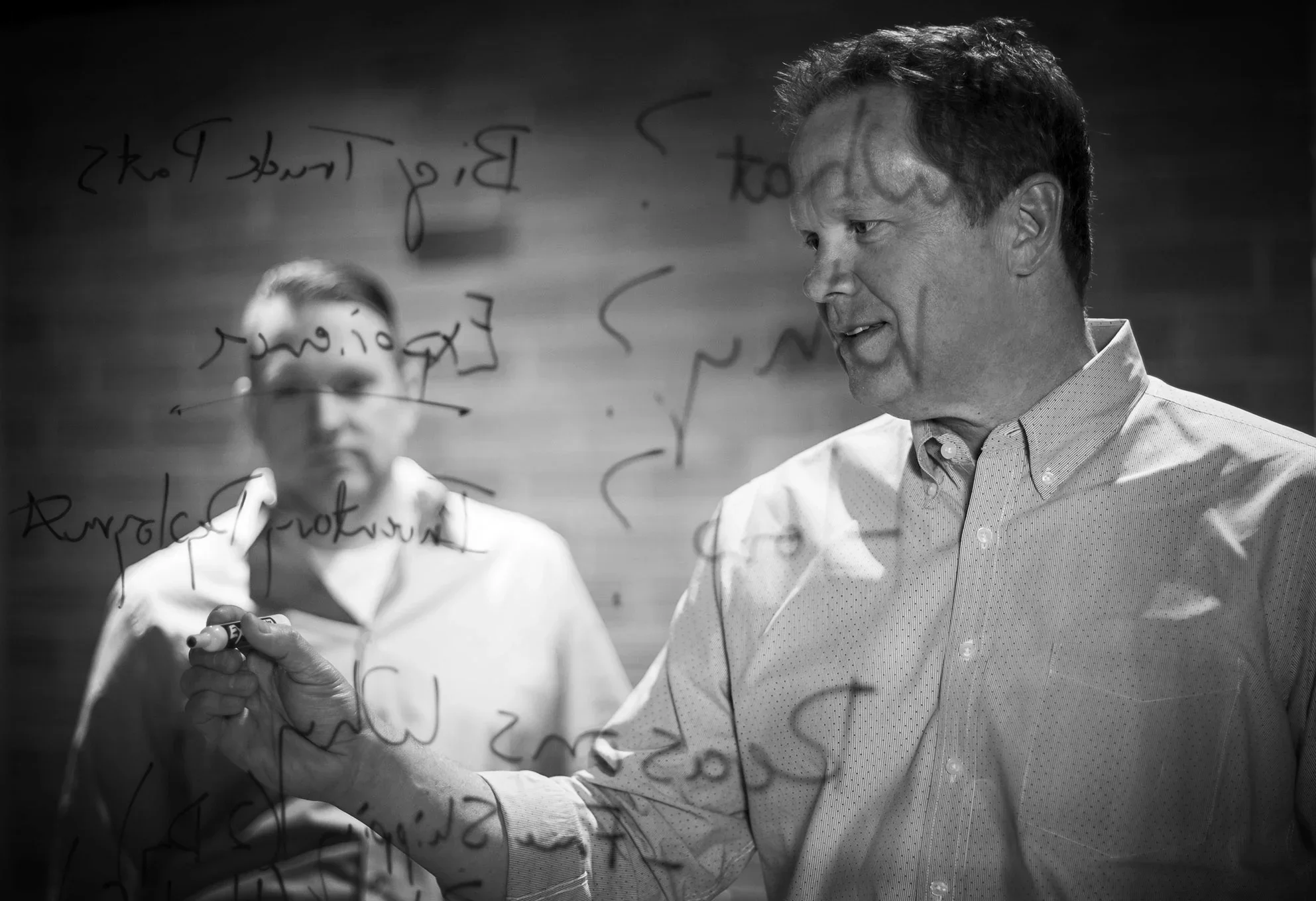 Two men working together on writing equations on a transparent glass surface, with one holding a marker and the other observing, both in formal shirts, in a professional setting.