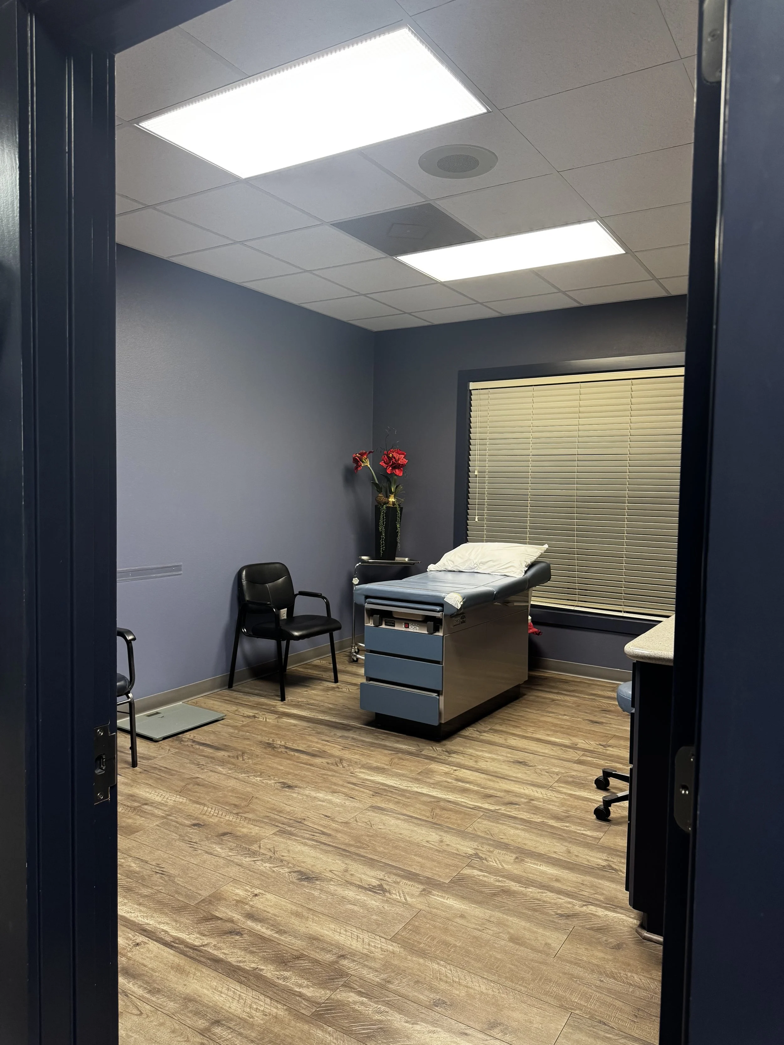 A medical examination room with gray walls, wooden floor, ceiling lights, a chair, an examination table with a pillow, a vase with red flowers, and a window with blinds.