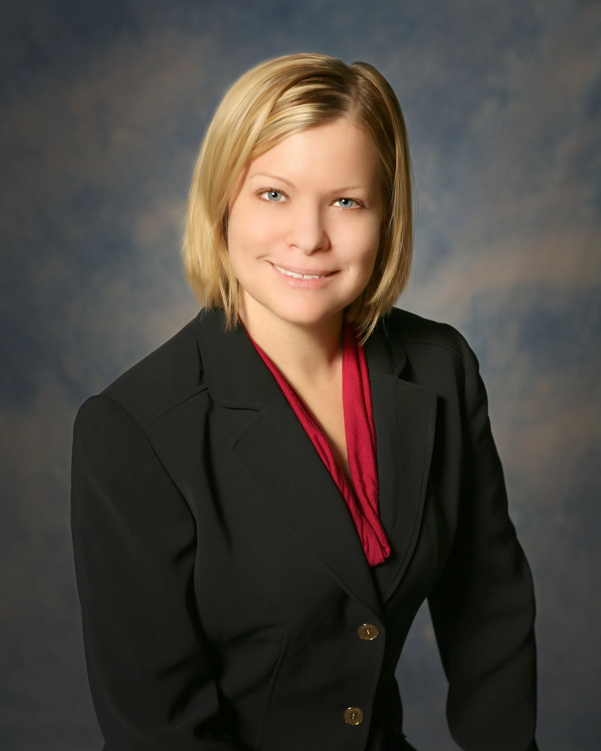 A woman with blonde hair, wearing a black blazer with gold buttons and a red blouse, smiling against a dark, blurred background.