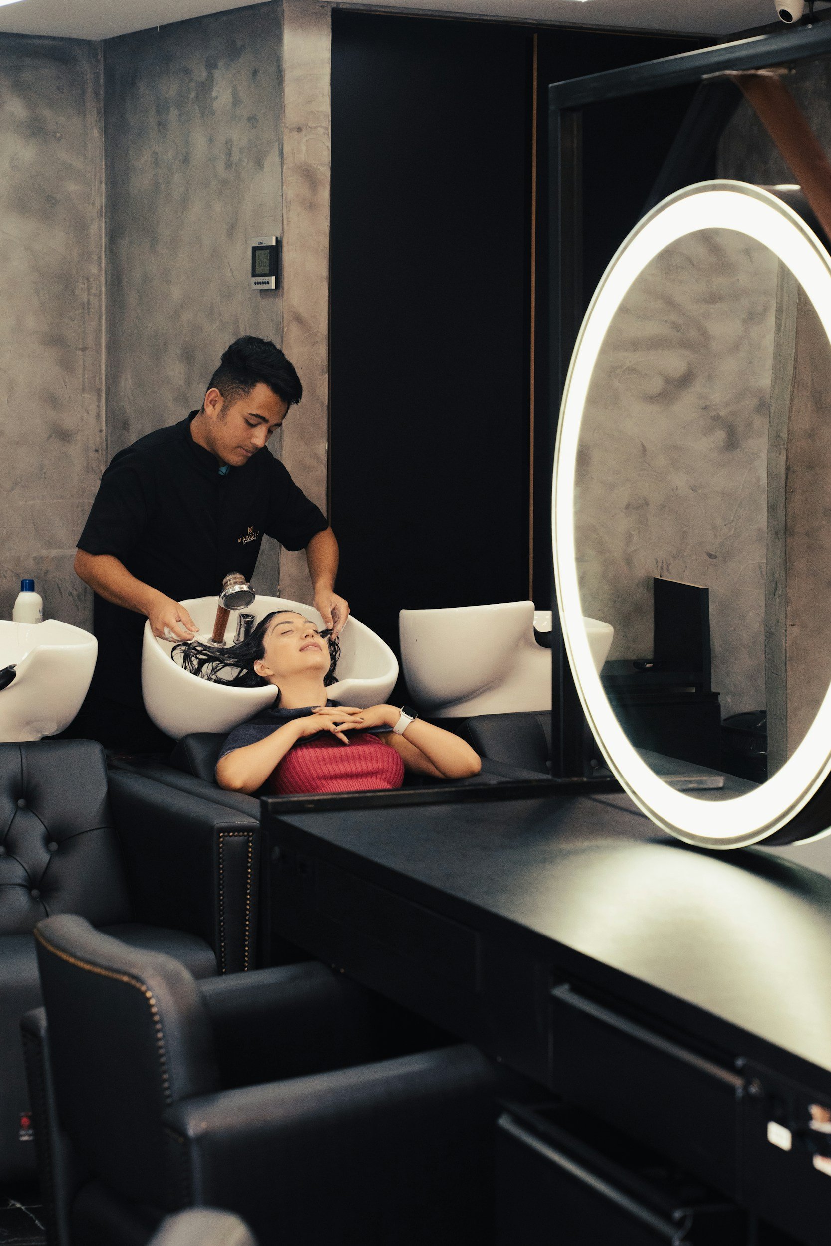 A woman with dark hair is reclining with her eyes closed in a styling sink while a hairstylist washes her hair. The scene is set in a modern salon with gray walls, a circular illuminated mirror, and black furniture.