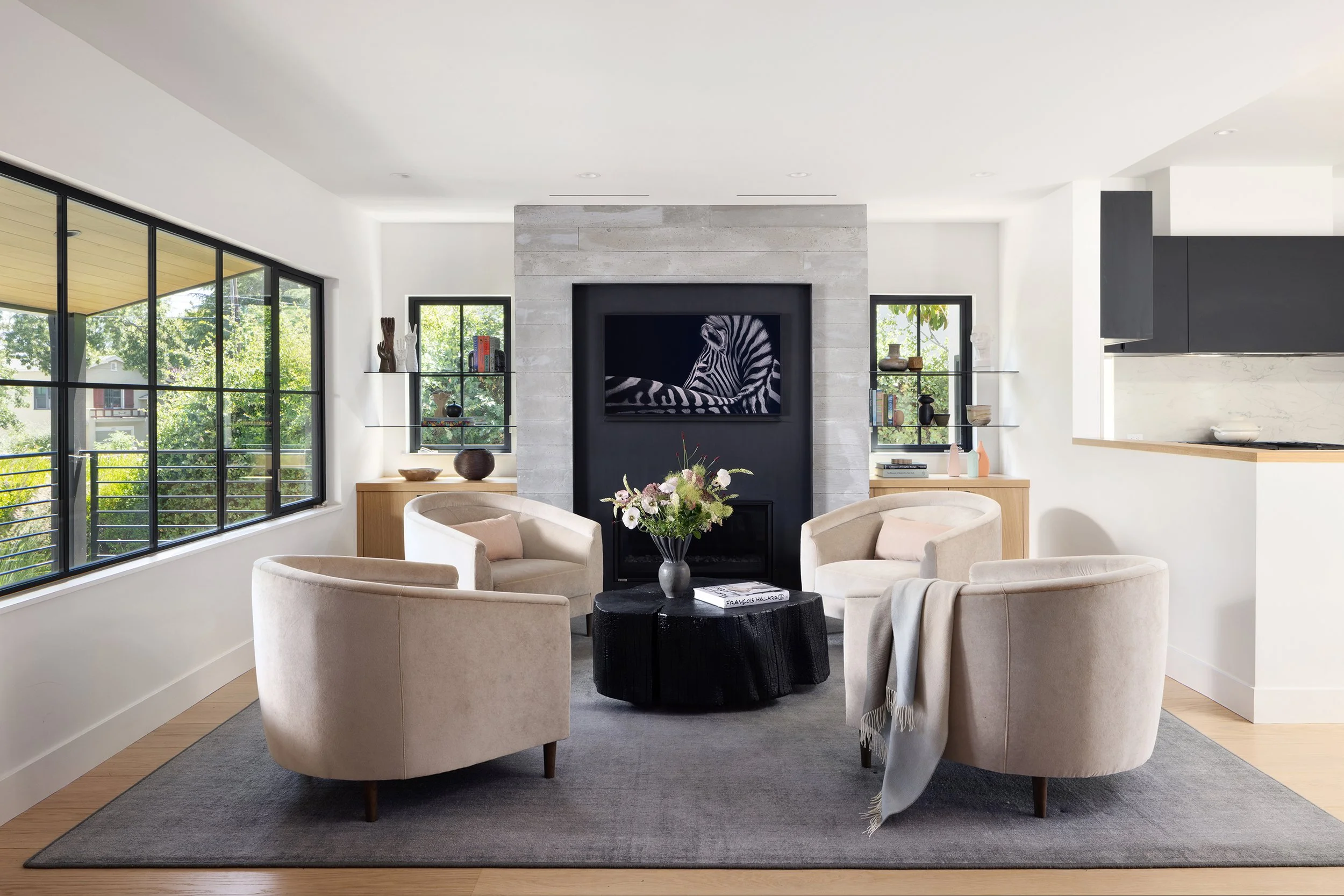 Modern living room with beige armchairs around a black coffee table, large windows, a TV mounted on a stone fireplace, and decorative items on shelves.