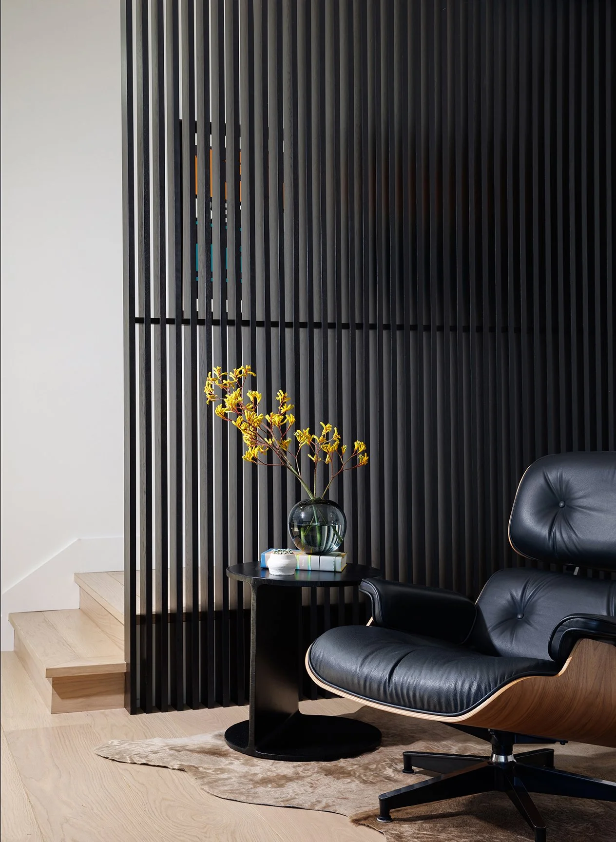Modern interior with black leather lounge chair, small black round side table with a glass vase holding yellow flowers, books, and a small container, black slatted wall, light wood staircase, and light wood floor with a beige rug.