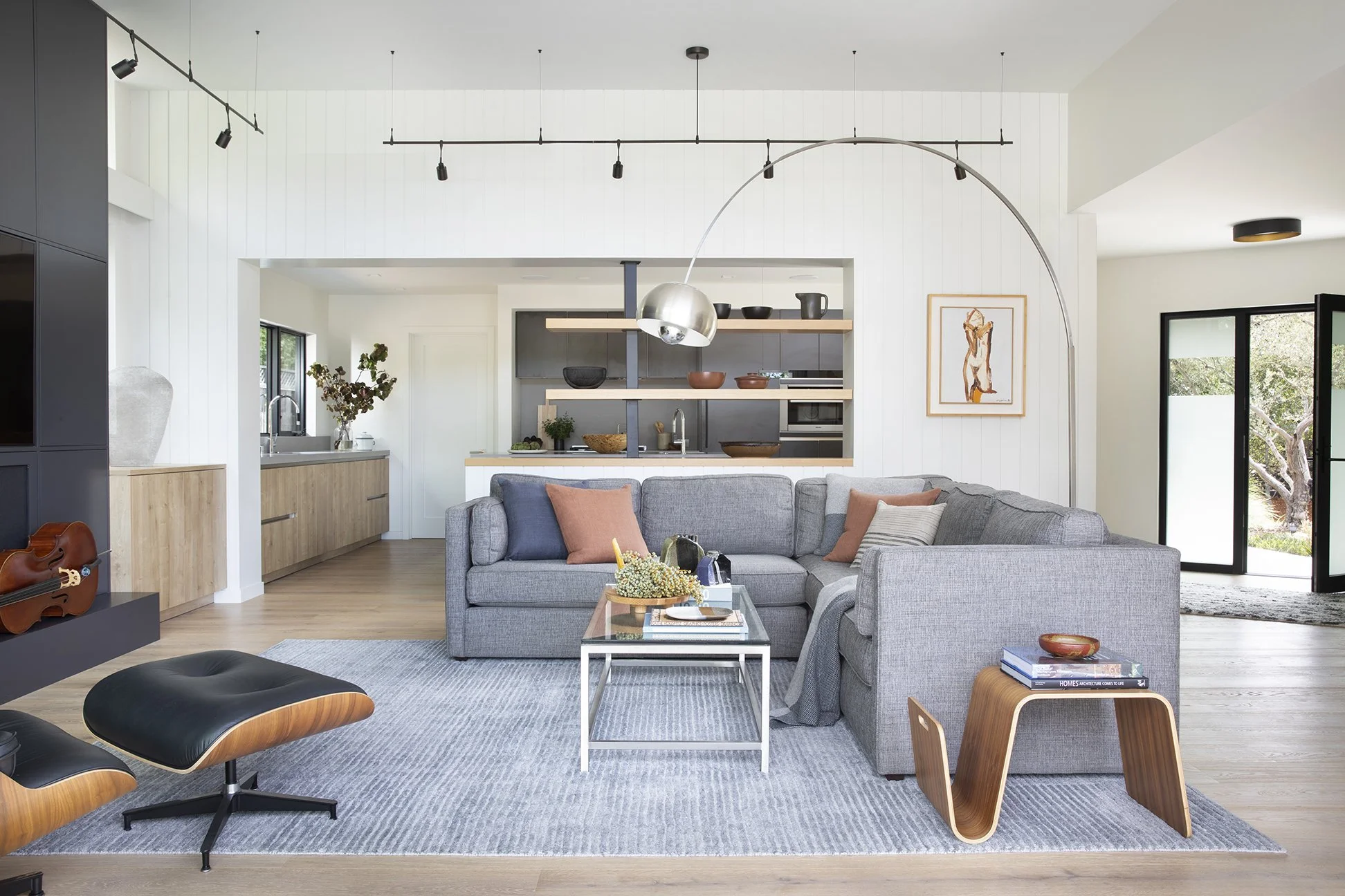 Modern living room with gray sectional sofa, glass coffee table, black lounge chair, and wooden furniture. Open kitchen in background with black and wood cabinets, artwork on wall, and sliding glass doors leading outside. Track lighting and pendant l