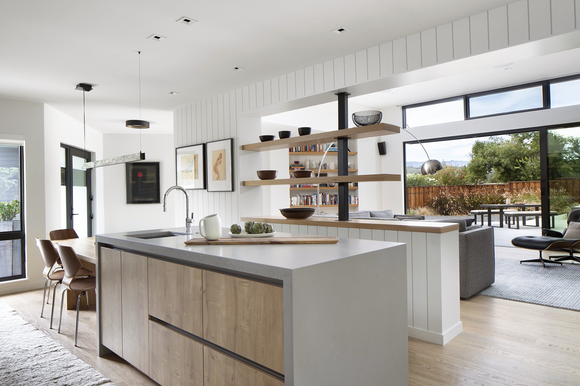 Open-concept living room and kitchen with large windows, white walls, wooden accents, modern furniture, and a backyard with greenery.