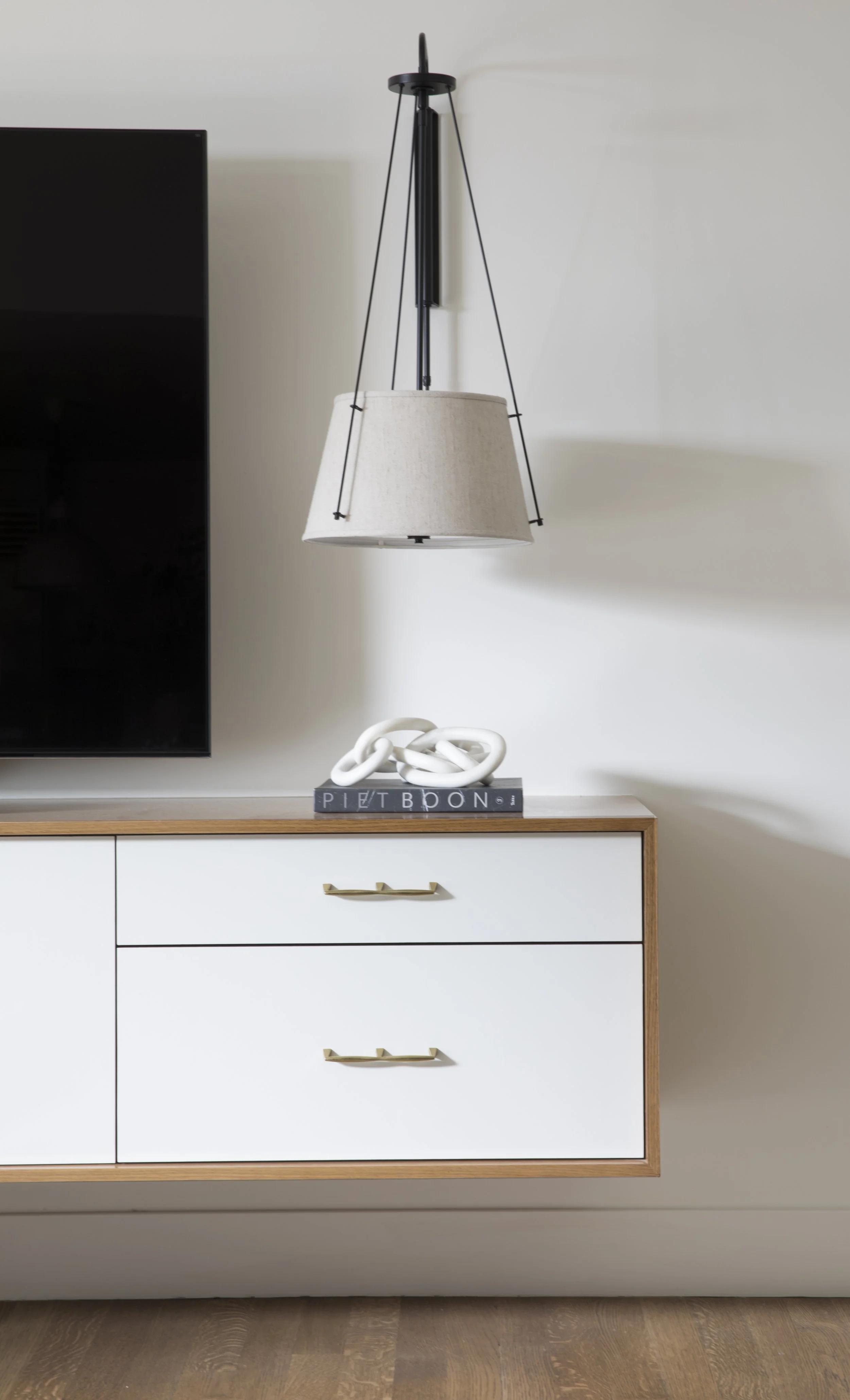 White dresser with three drawers, gold handles, and a wooden frame, topped with a decorative white sculpture and a book titled 'PIET BOON'. A modern black and white lamp hangs above and to the right of the dresser, with a light beige lampshade, again