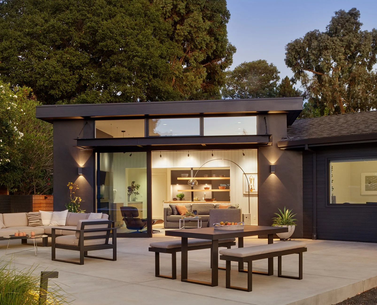 Modern house with large glass sliding doors open to outdoor patio with seating area and interior living room visible inside, surrounded by tall trees.