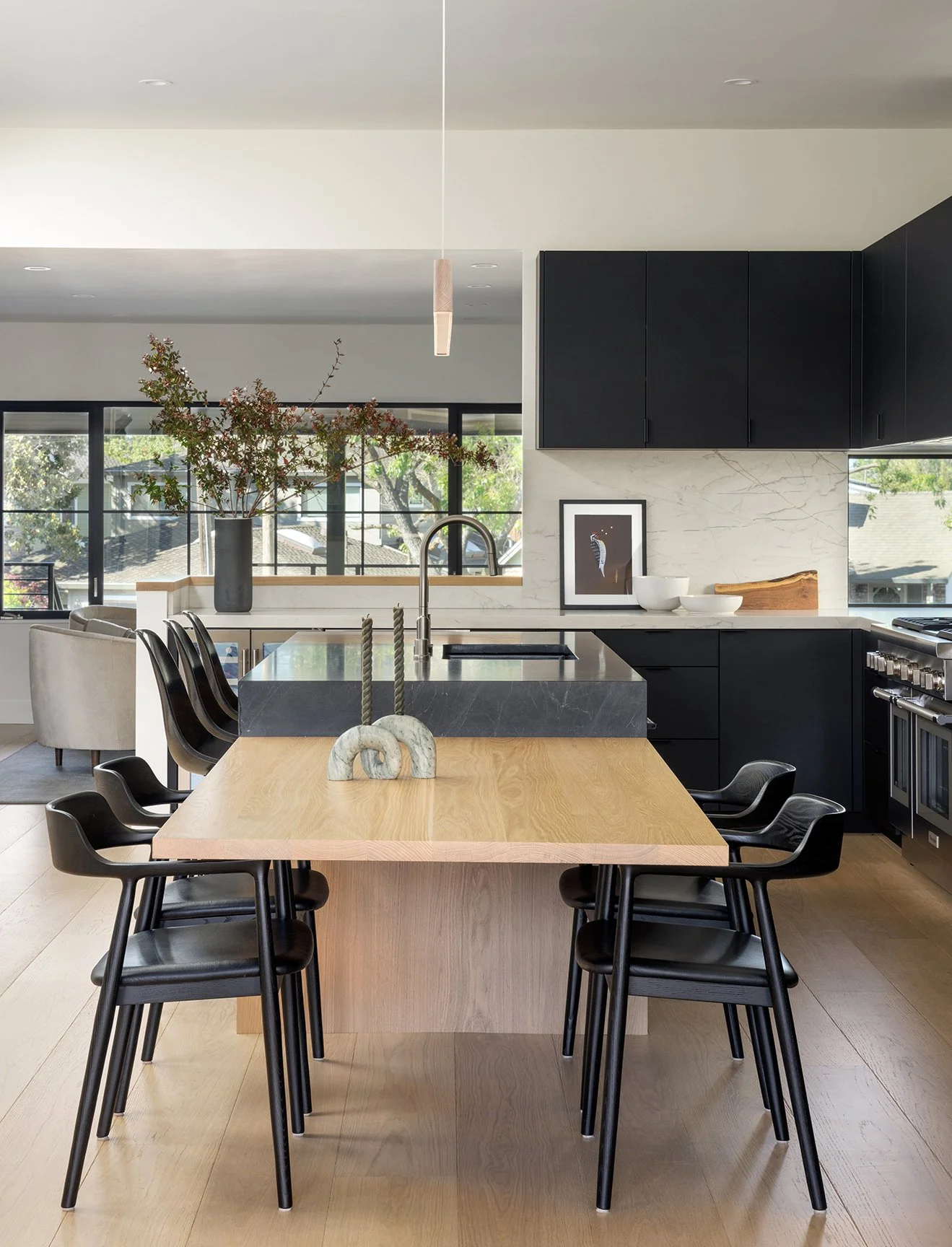 Modern kitchen with a wooden dining table and black chairs, black cabinetry, marble backsplash, large windows, and a decorative vase with branches.