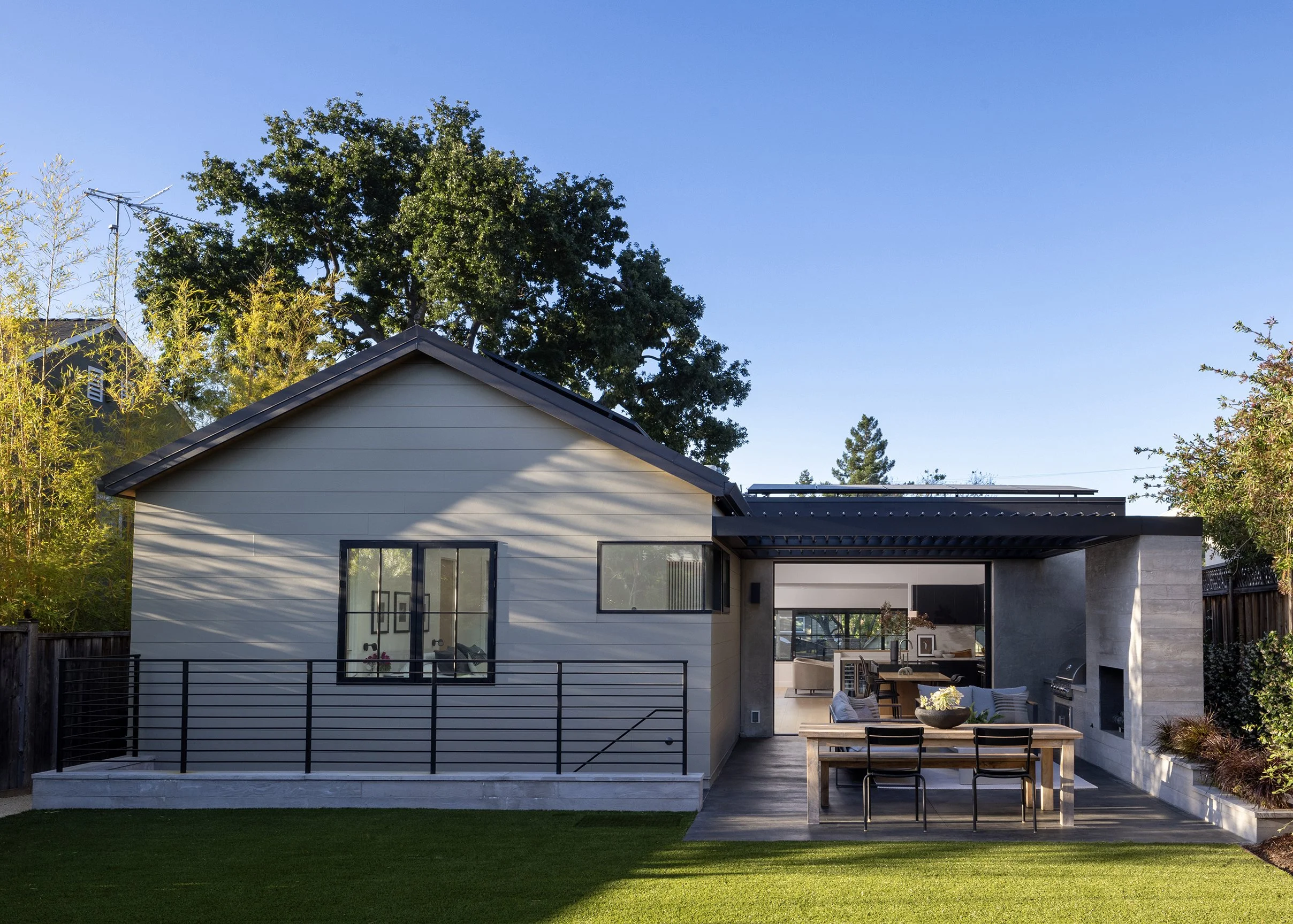 Modern backyard with a patio, outdoor seating, a table with a bowl, and a house with large windows, surrounded by trees and greenery under a clear blue sky.