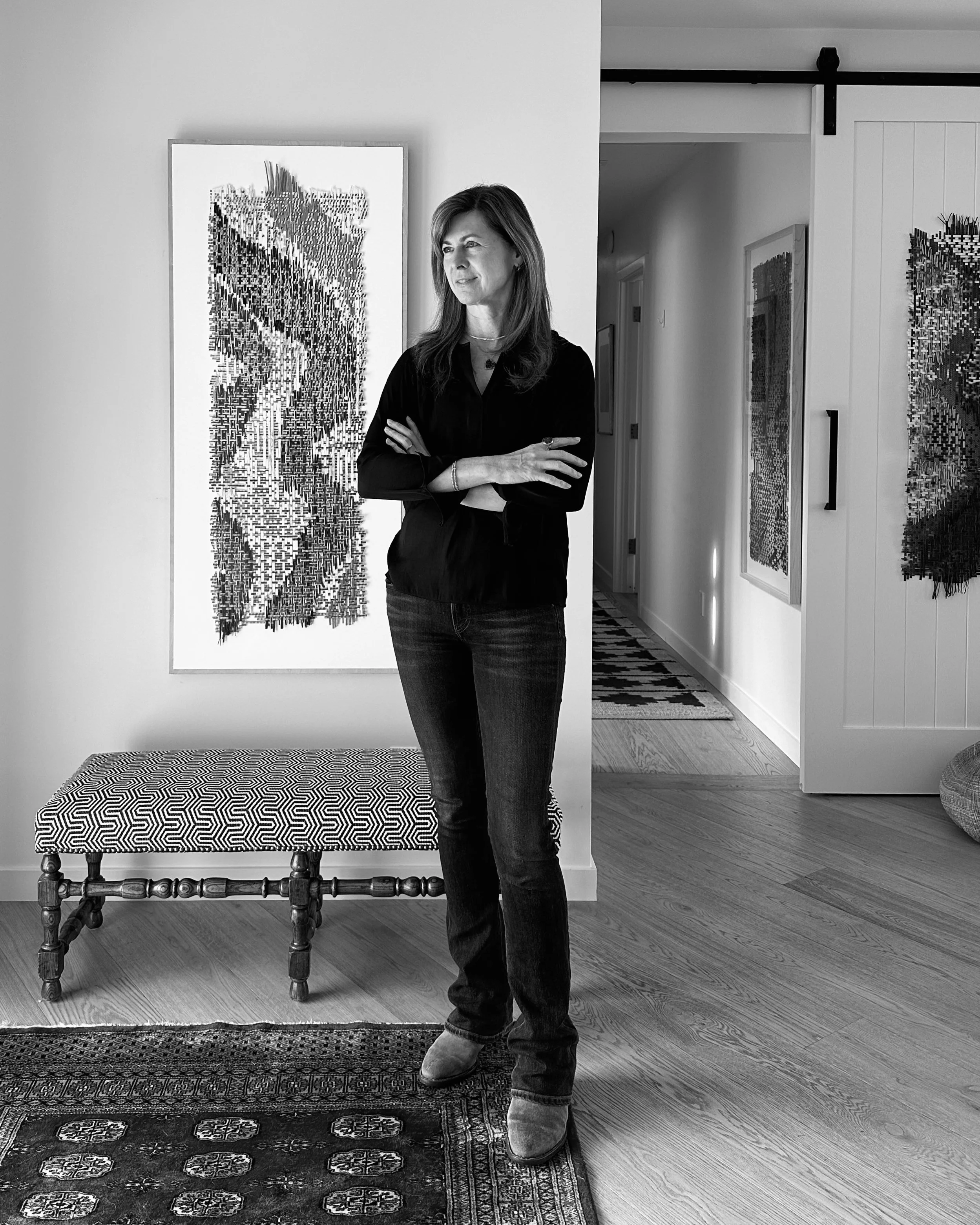 A woman with long hair wearing a dark top and jeans, standing with arms crossed, in an indoor space that appears to be an art gallery. She is near framed artwork on the wall, with a patterned carpet and wooden floors visible.
