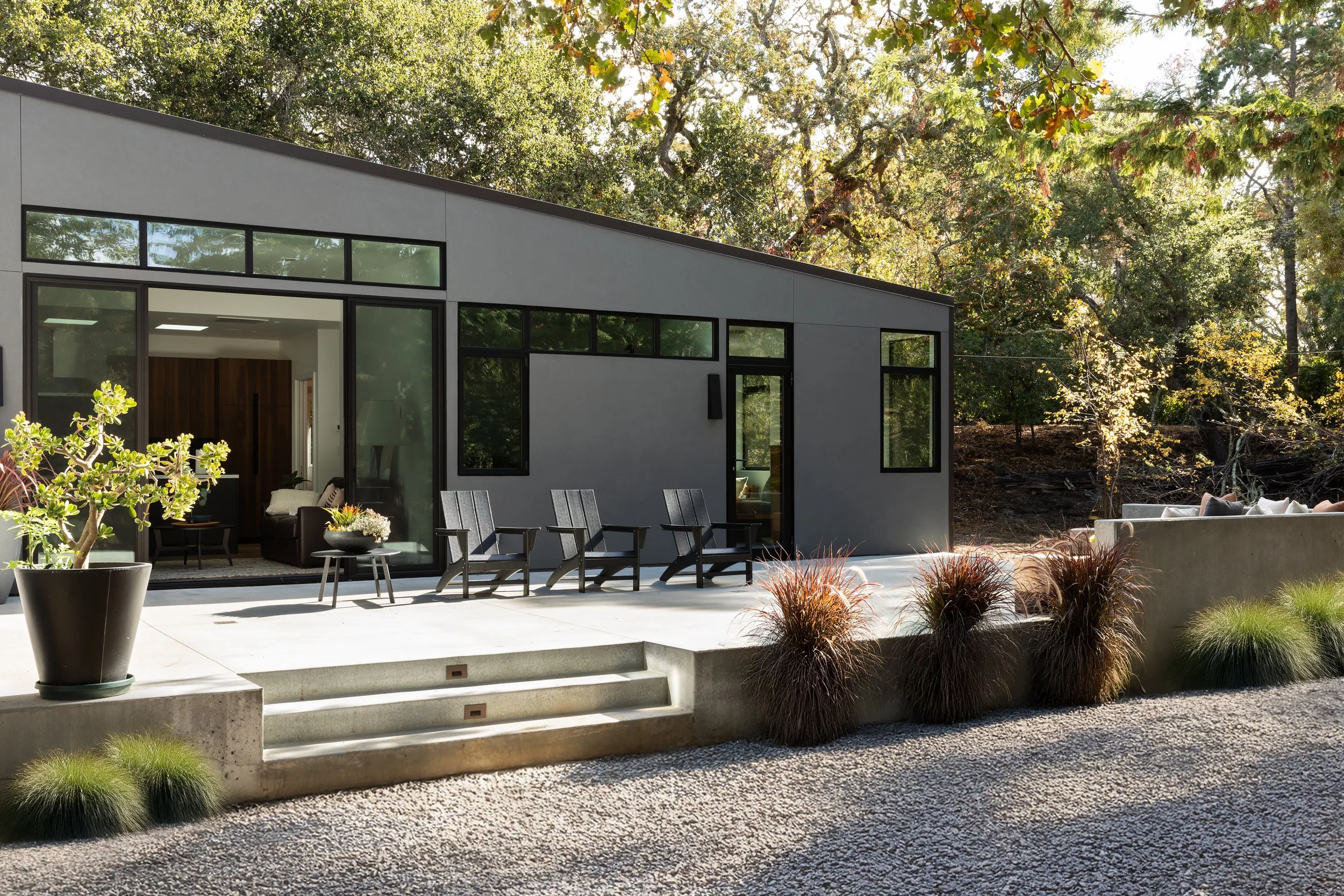 Modern house with large glass windows and sliding doors, outdoor patio with chairs, plants, and steps, surrounded by trees and landscaped yard, during the daytime.