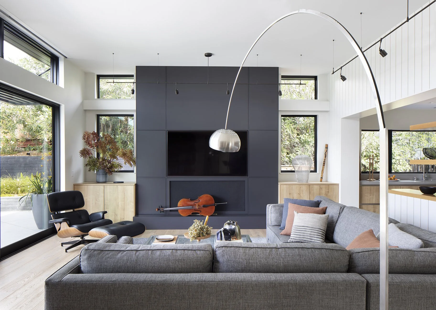Modern living room with large windows, gray sectional sofa with pillows, black lounge chair, wooden cabinets, a large flat-screen TV, a cello on the black entertainment wall, and contemporary decor.
