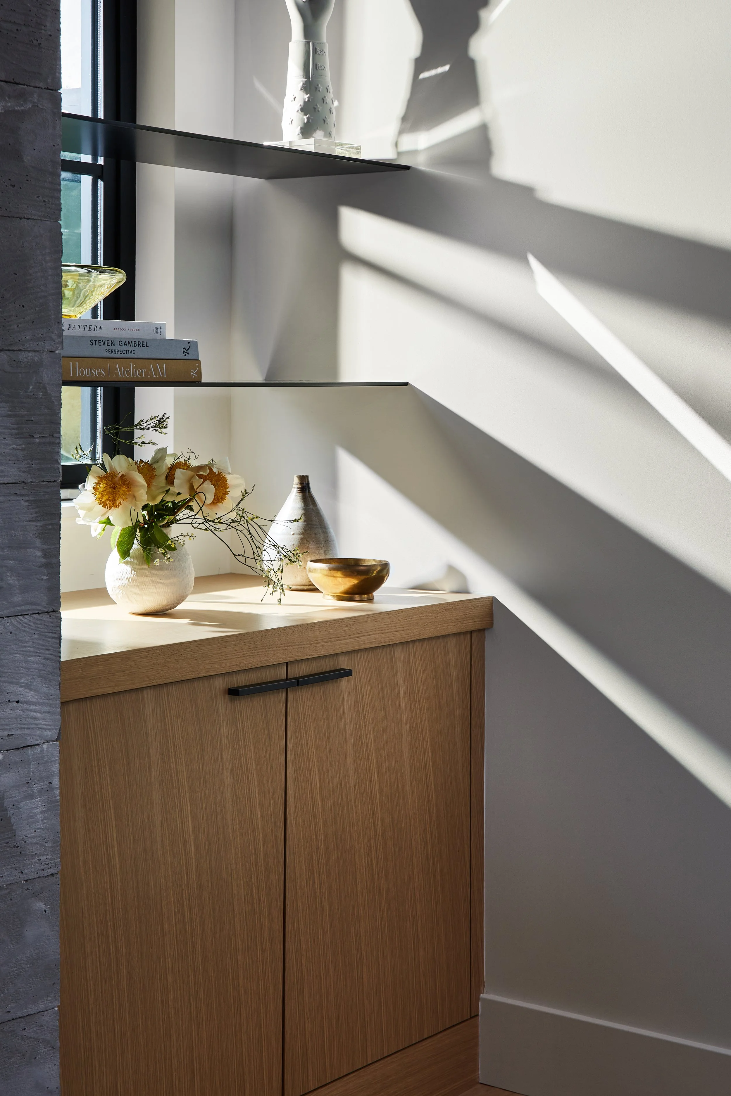 A wooden cabinet with a flat top surface, decorated with a white vase holding flowers, small bowls, and decorative objects, illuminated by sunlight and casting shadows on the wall behind.