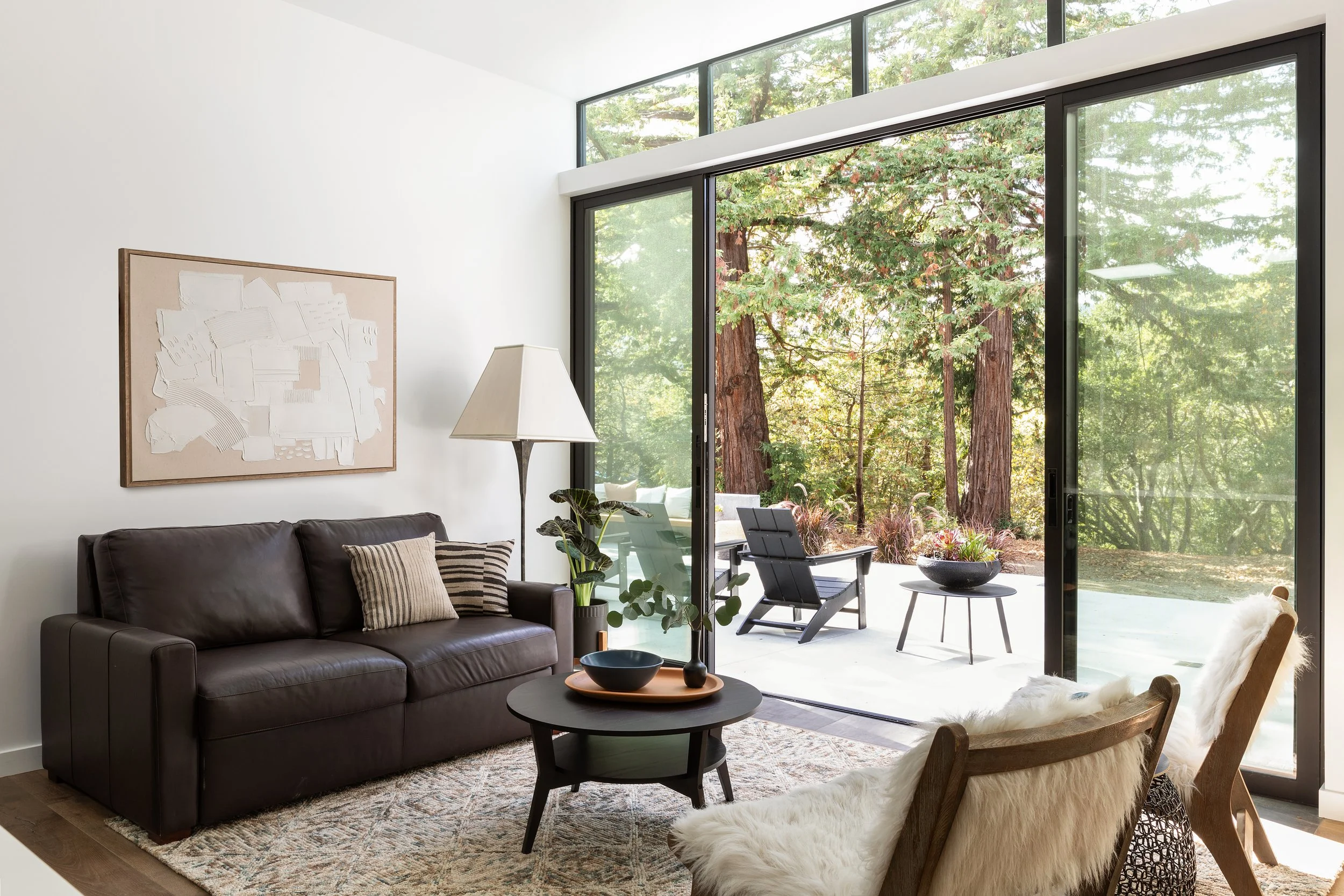 Modern living room with a black leather sofa, a round coffee table, and a wood chair with a white furry cushion, large floor-to-ceiling windows showing outdoor trees, and a white wall with abstract art.