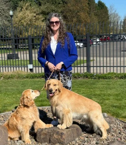 A woman with long wavy hair wearing sunglasses, a blue blazer, and a white top standing outdoors on grass, holding two Golden Retrievers, one puppy and one adult, near a metal fence with trees and parked cars in the background.