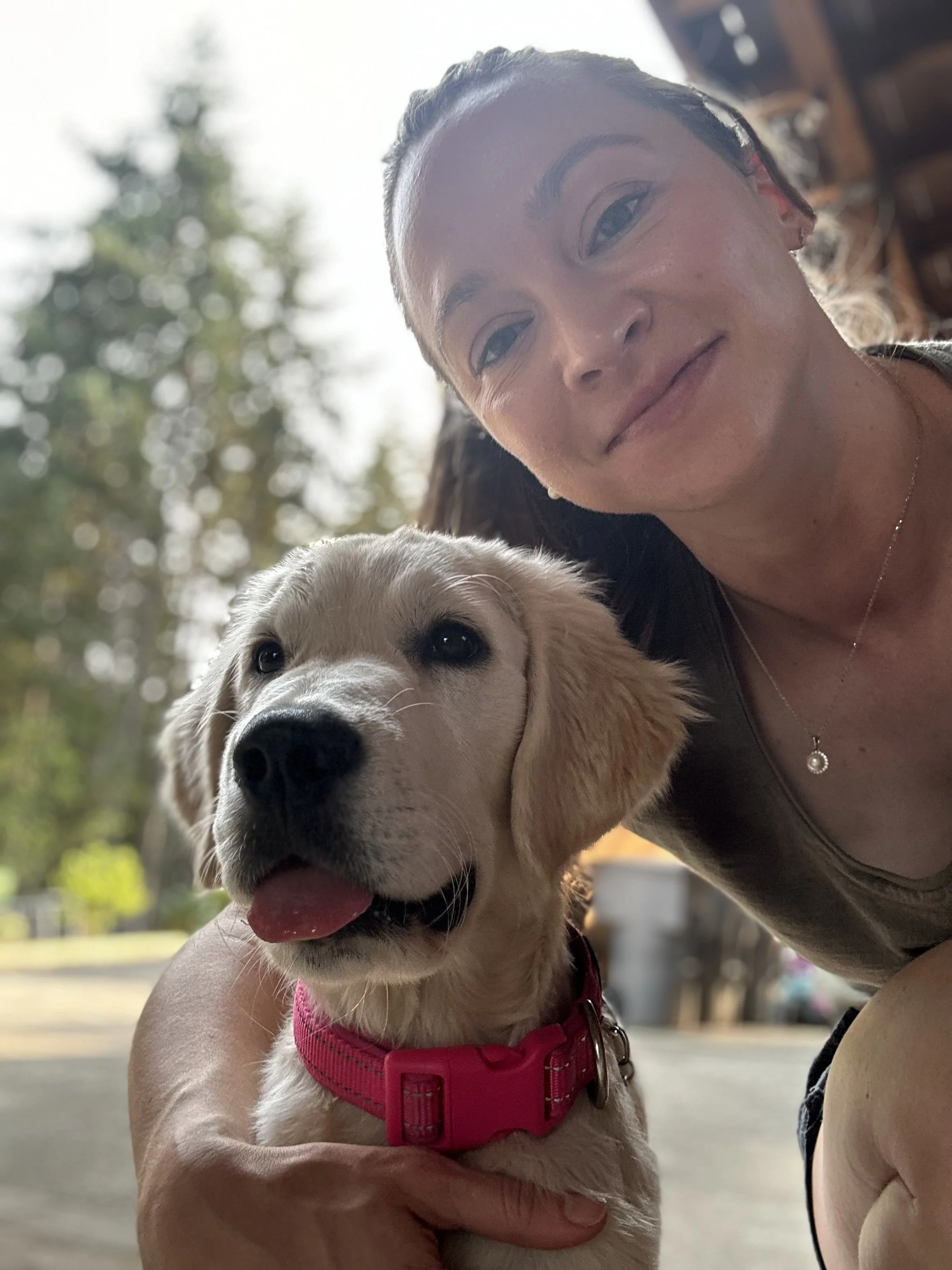A woman with her hair pulled back taking a selfie with a happy puppy, both outdoors with blurred trees in the background.