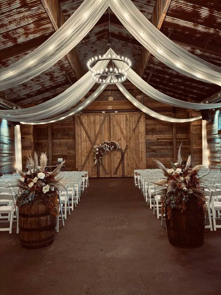 Wedding ceremony setup inside a rustic barn with wood walls and ceiling, decorated with flowing white fabric, floral arrangements in wooden barrels, and a heart-shaped floral arch at the altar.
