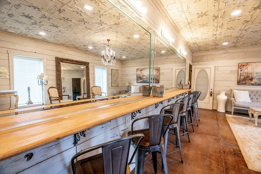 Interior of a rustic-style room with a long wooden bar counter, metal chairs, a large mirror, chandelier, and paintings on wood-paneled walls.