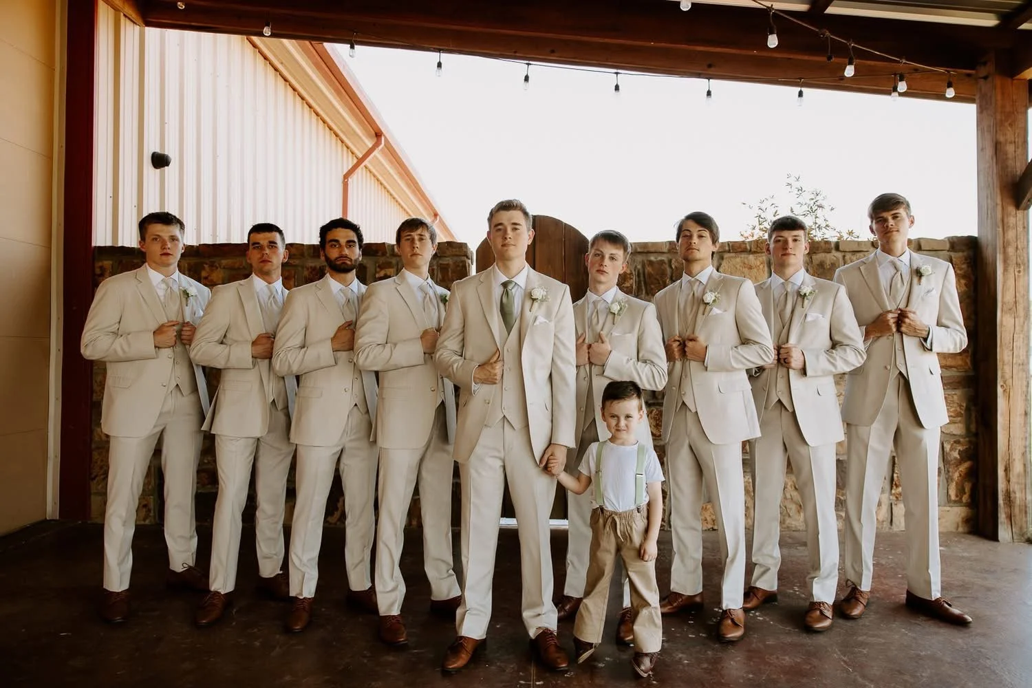 A group of nine men and one young boy dressed in white suits with boutonnières, standing in front of a rustic brick and wood backdrop, with the young boy holding hands with the man in the center, at an outdoor wedding or formal event.