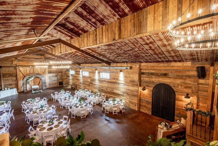 A rustic wedding reception hall with wooden walls and ceiling, round tables with white tablecloths and gold chargers, floral centerpieces, and a decorative arch at the front.