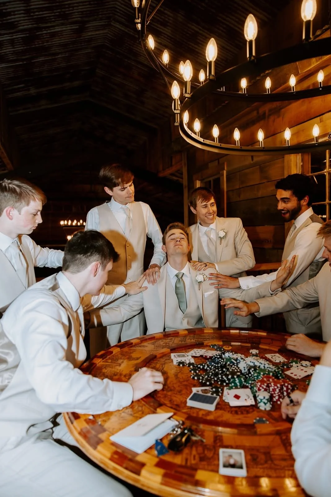 A group of men in beige suits celebrating around a poker table with chips, cards, and a photo, inside a rustic wooden building with a large chandelier.