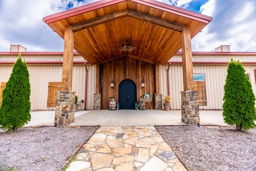 Front entrance of a rustic barn-style building with a large covered porch, stone pillars, and decorative items near the door, flanked by two tall evergreen bushes.