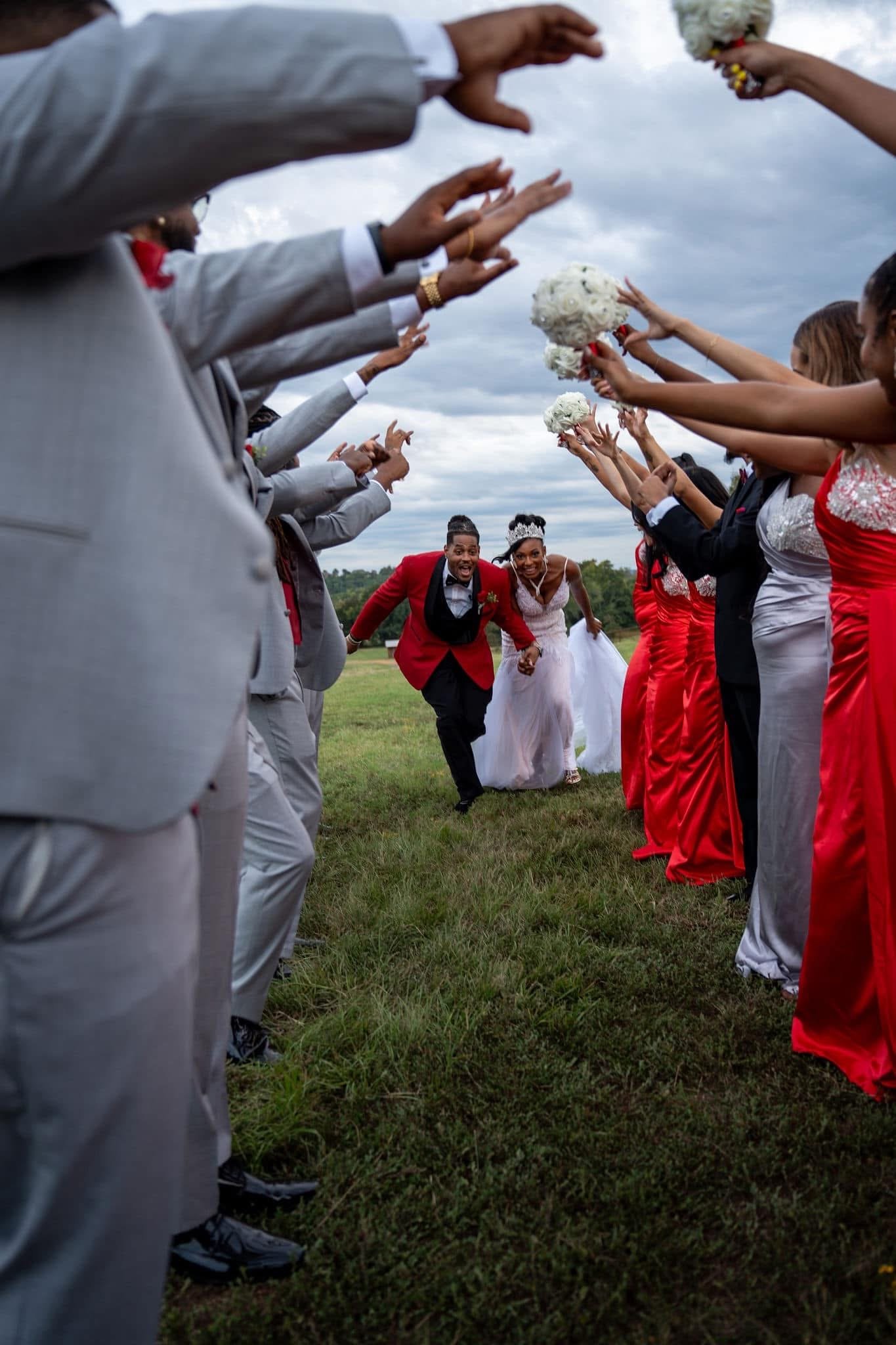 A bride and groom walking through a tunnel created by wedding guests holding bouquets, with guests dressed in gray and red suits and dresses, outdoors on a grassy field with overcast sky.