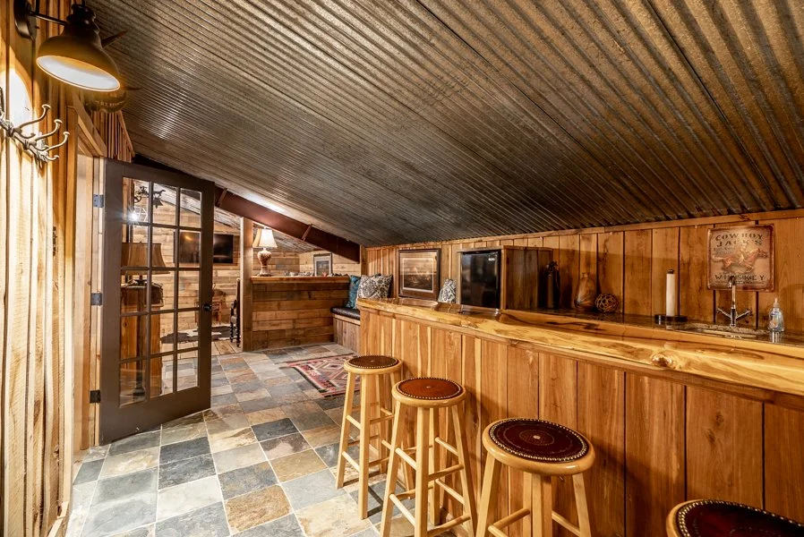 Cozy attic bar with wooden paneling, a slate tile floor, three stools, a bar counter, and warm lighting.