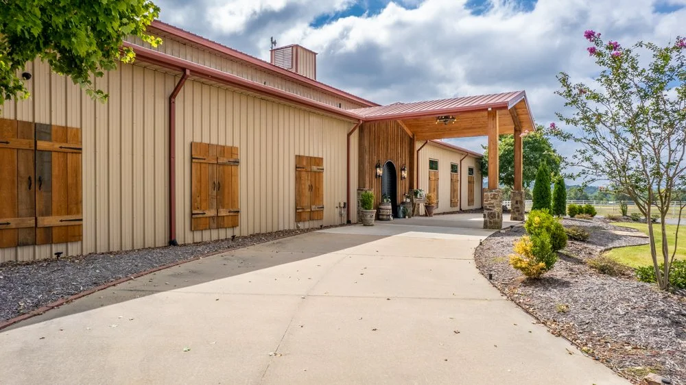 Exterior view of a ranch-style building with a red metal roof, wooden shutters, and a covered porch with stone columns, surrounded by landscaped greenery and trees.