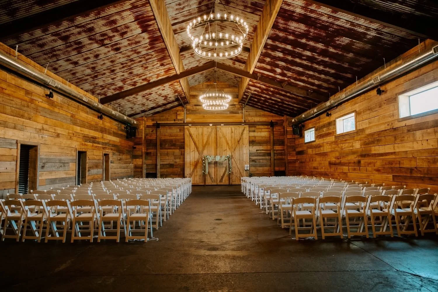 Indoor wooden wedding venue with rows of white chairs facing a large wooden barn door, decorated with greenery, and chandelier lighting.