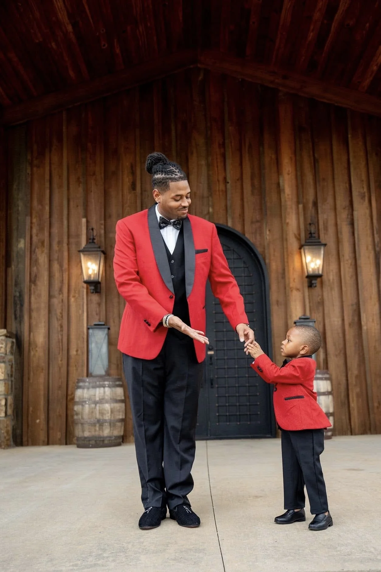 A man and a young boy, both dressed in matching red and black suits, are holding hands and facing each other. They are standing outside in front of a wooden building with lanterns on the wall and barrels on each side.