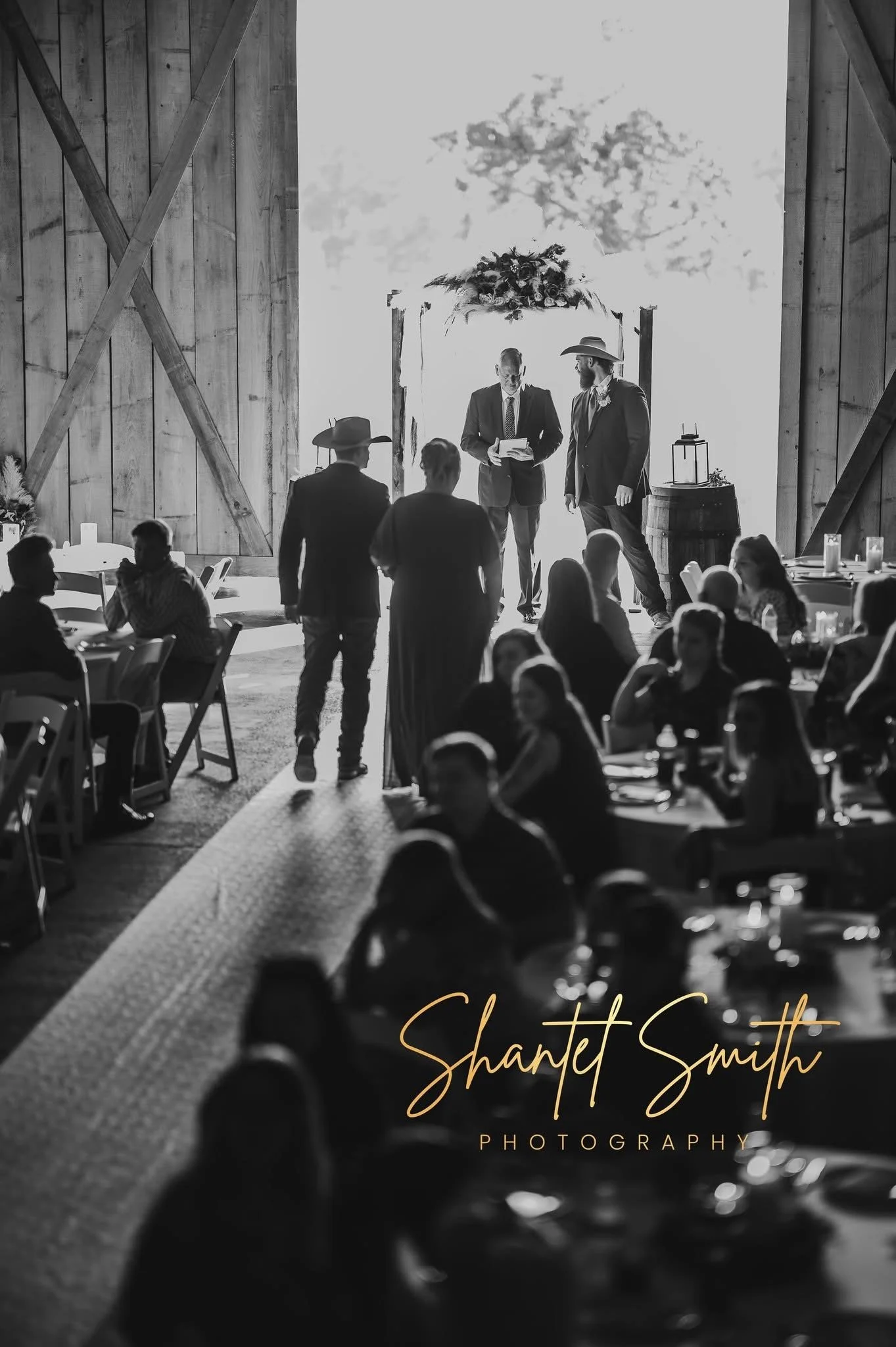 Black and white photo of a wedding ceremony in a barn with floral arch, officiant, and groom, guests seated at tables, and the photographer's watermark saying 'Sharnett Smith Photography' at the bottom.