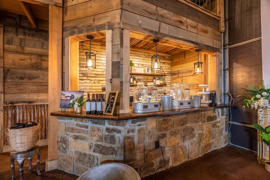 Indoor wooden bar with stone base, lamp lighting, water dispensers, glass jars, wine bottles, framed photos, and decorative plants.