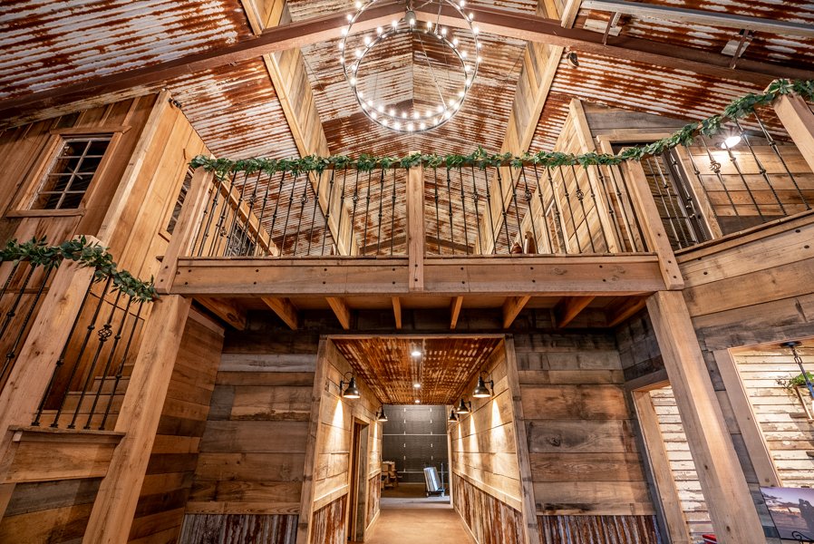 Interior of a rustic barn with wooden walls and ceiling, featuring a second-floor balcony decorated with green garland. A circular chandelier hangs from the ceiling, illuminating the space.