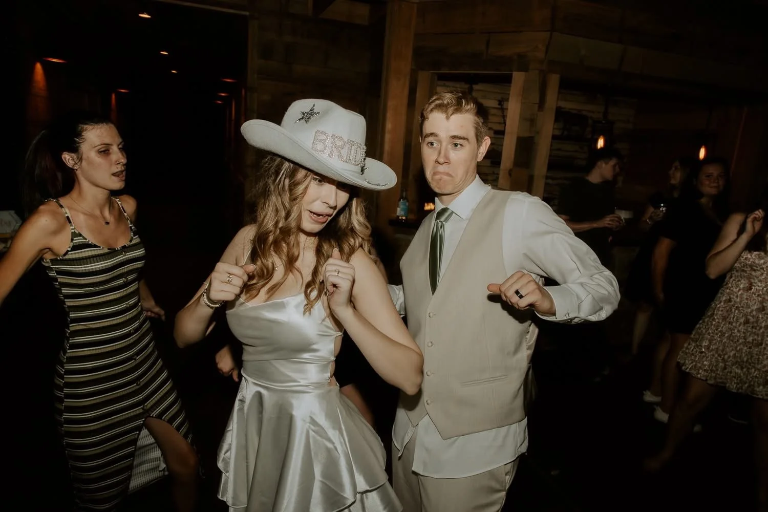 People dancing at a wedding reception, with a woman wearing a corset-style white dress and a large hat that says 'BRIDE', and a man in a beige vest and white shirt.