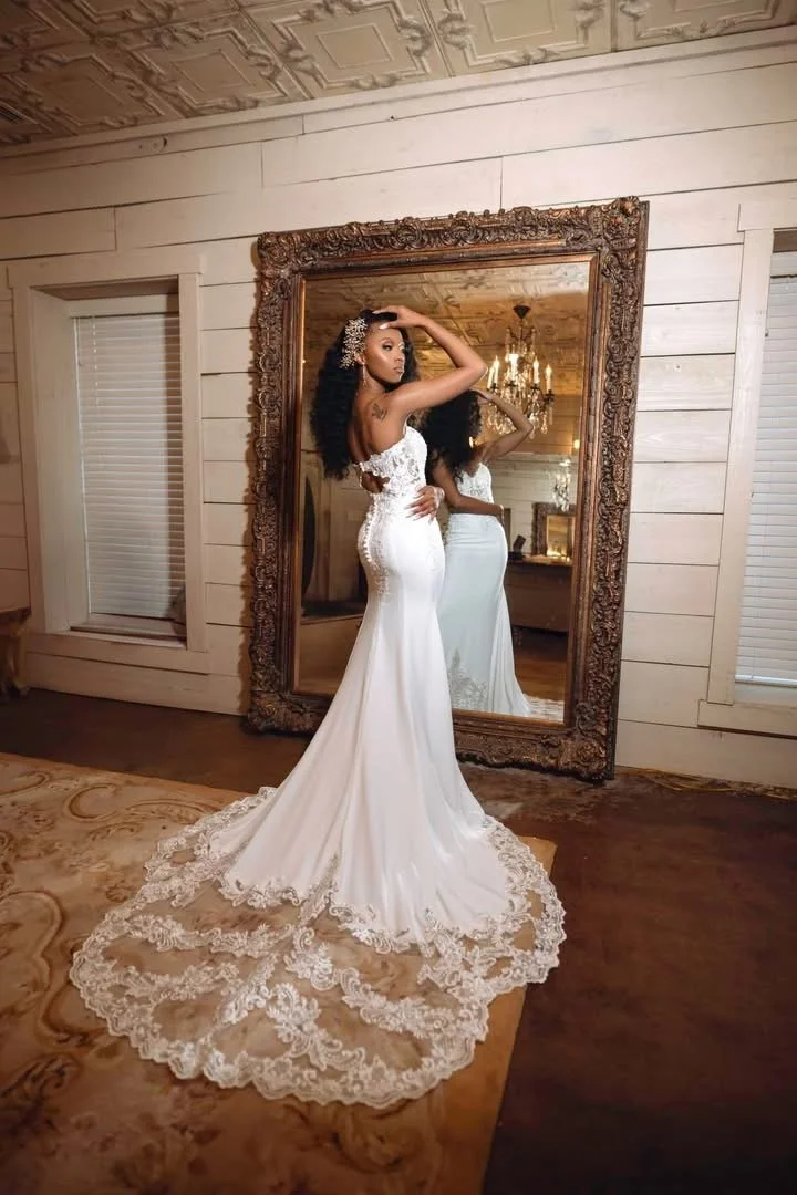 A woman in a white wedding dress with lace details standing in front of a large ornate mirror in a room with wooden walls and a chandelier visible through the mirror.