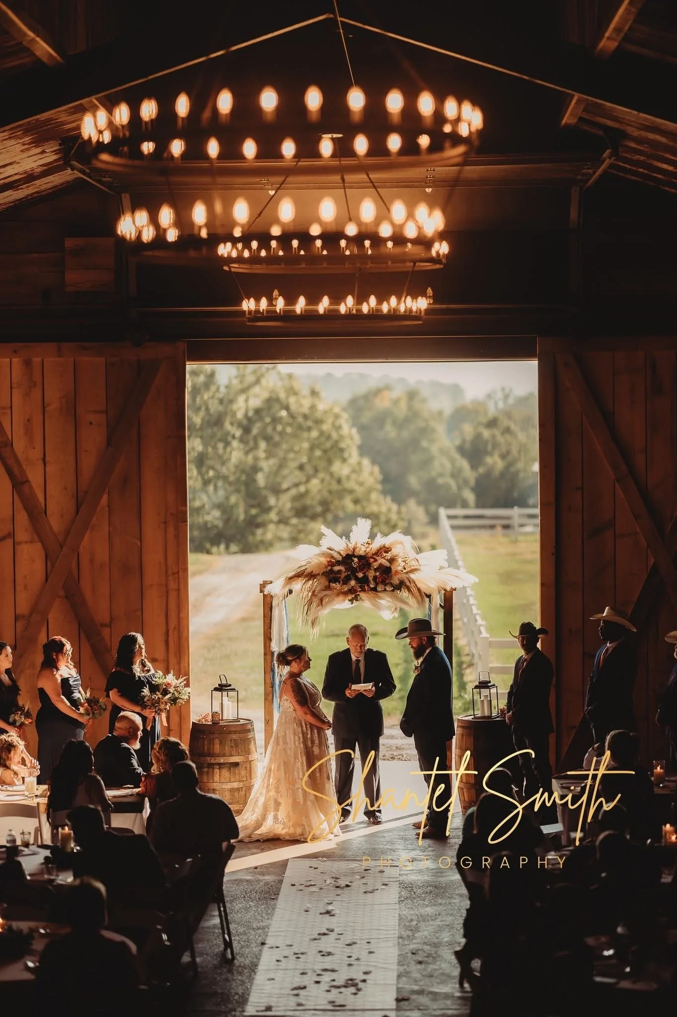 Wedding ceremony inside a rustic barn, with a couple standing before an officiant, surrounded by wedding guests. The barn features large wooden doors, a high ceiling with a chandelier, and a scenic outdoor view.