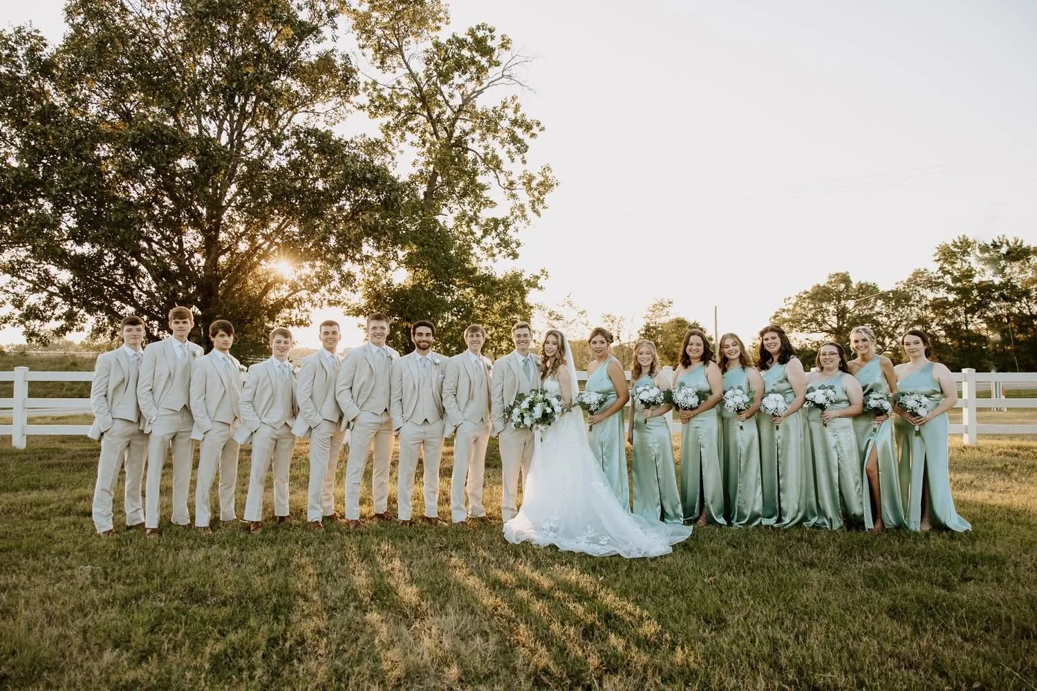A group of wedding party members, including the bride and groom, standing outdoors on grass during sunset, with trees and a white fence in the background.