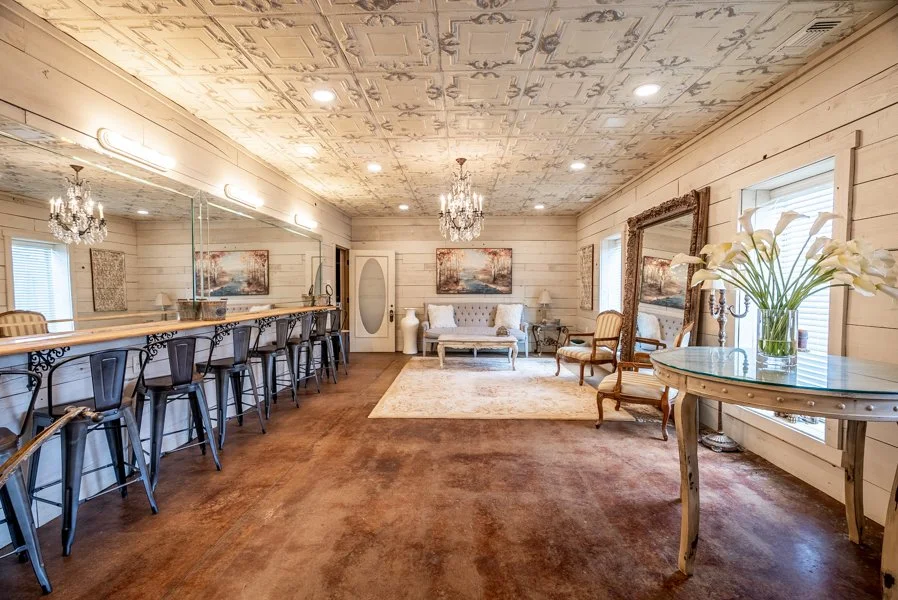 A vintage-style lounge with chandeliers, artwork, and wooden paneling. It features a bar area with high metal chairs, a white sofa, armchairs, a large mirror, and a round table with flowers.