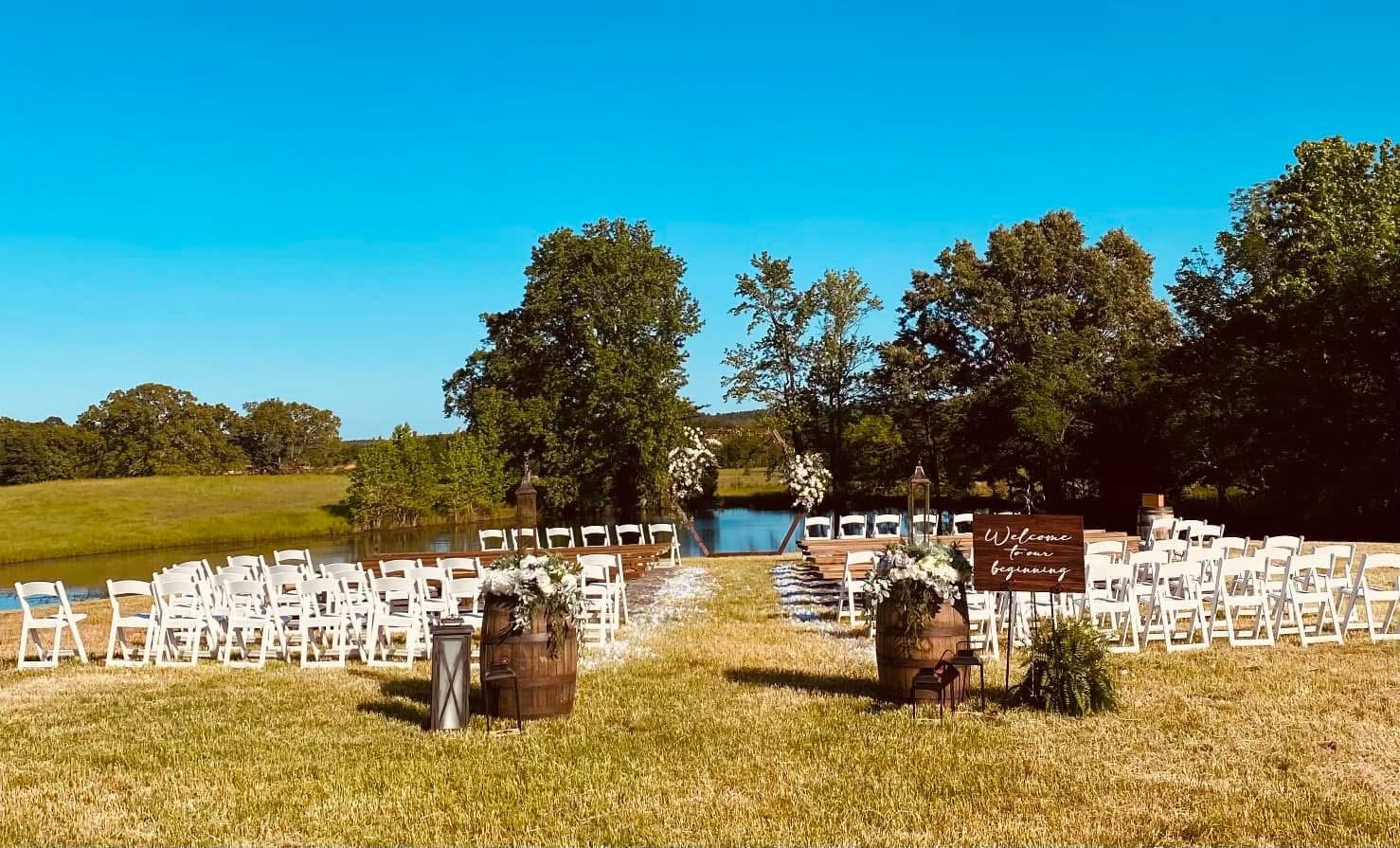 Outdoor wedding setup on a grassy field by a lake with white folding chairs arranged in rows, decorated with floral arrangements, a sign reading 'Welcome to our beginning,' and a central aisle leading to a water view with trees in the background.